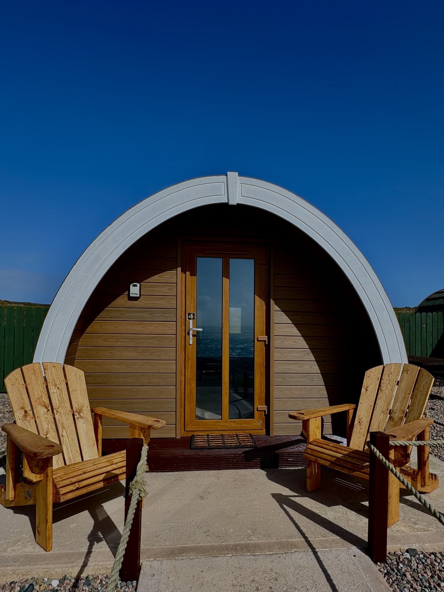 A glamping pod exterior with timber cladding, an arched grey roof and two wooden chairs on the patio under deep blue sky.